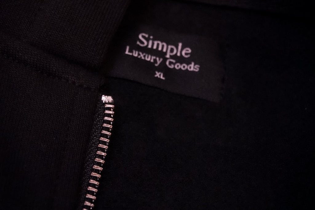 Close-up of a black garment showing a sewn-in  woven label that reads 'Simple Luxury Goods' and 'XL', with a silver zipper running vertically along the left side.