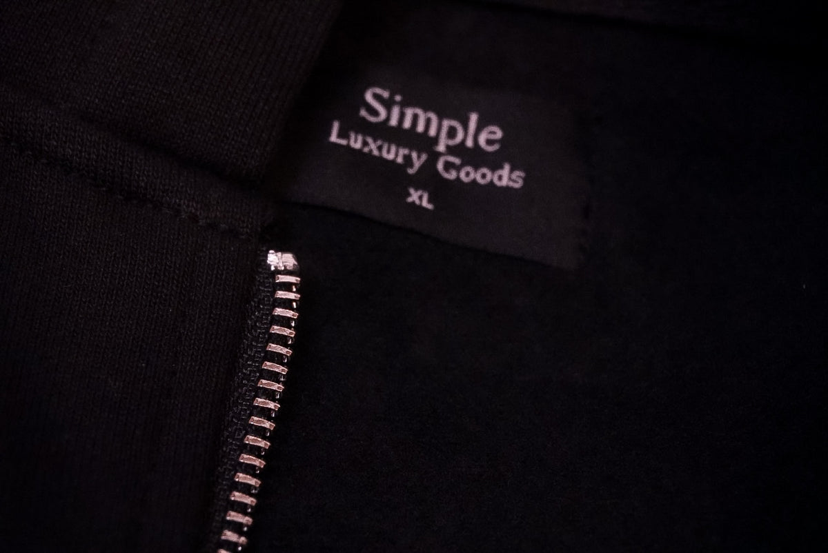 Close-up of a black garment showing a sewn-in  woven label that reads 'Simple Luxury Goods' and 'XL', with a silver zipper running vertically along the left side.