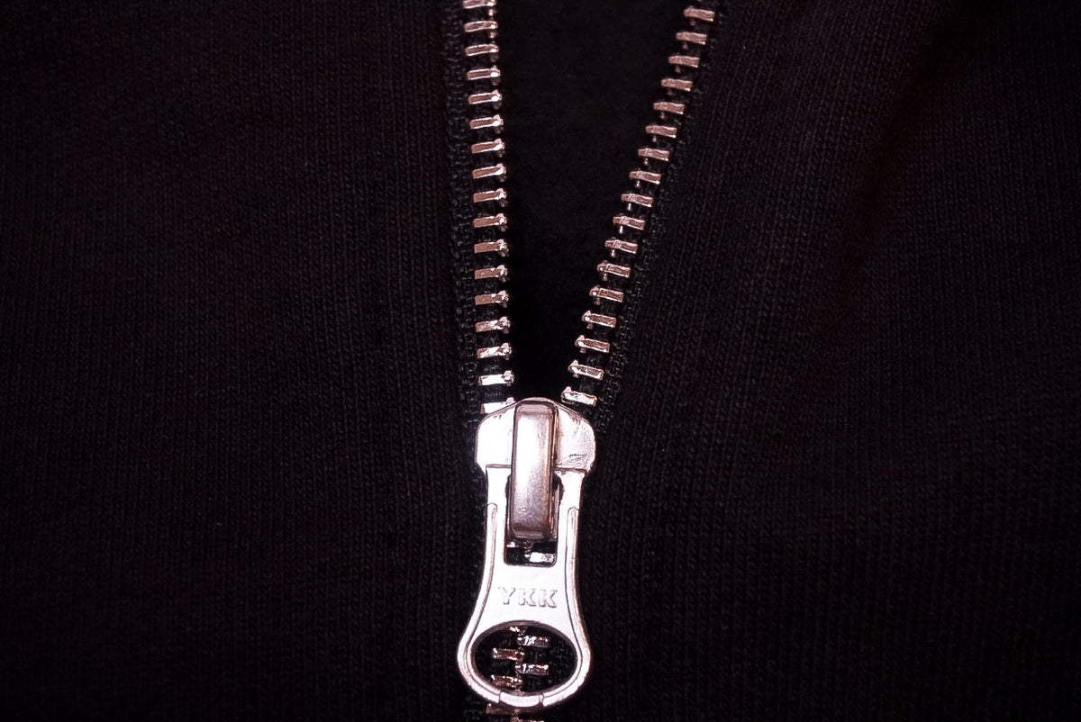 A close-up of a silver metal zipper pulled open on a black fabric garment, showing the teeth of the zipper and a silver pull tab against the black fabric.
