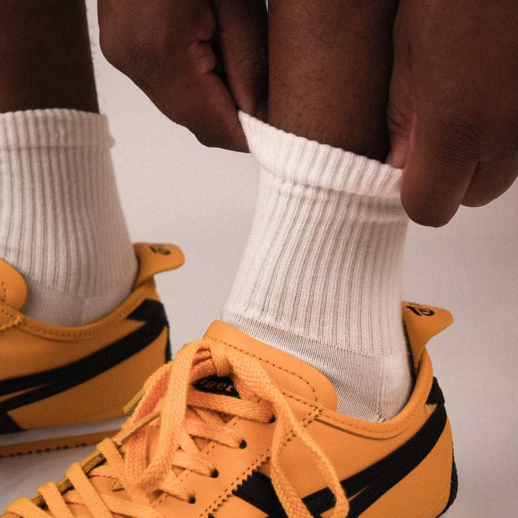 A close-up of a person's hands pulling up a white ribbed quarter sock on their ankle, with a yellow and black sneaker visible below.