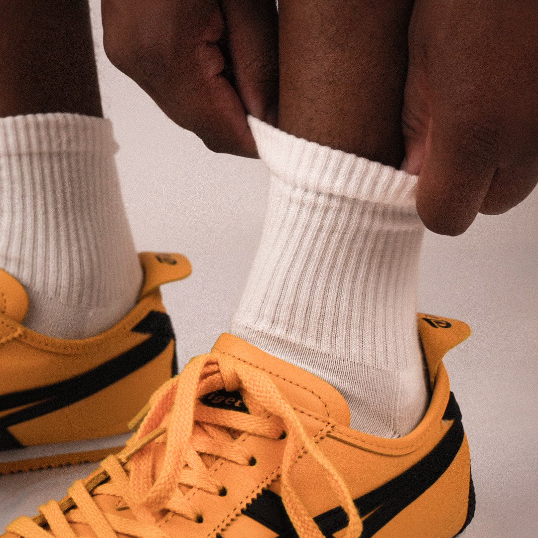 A close-up of a person's hands pulling up a white ribbed quarter sock on their ankle, with a yellow and black sneaker visible below.