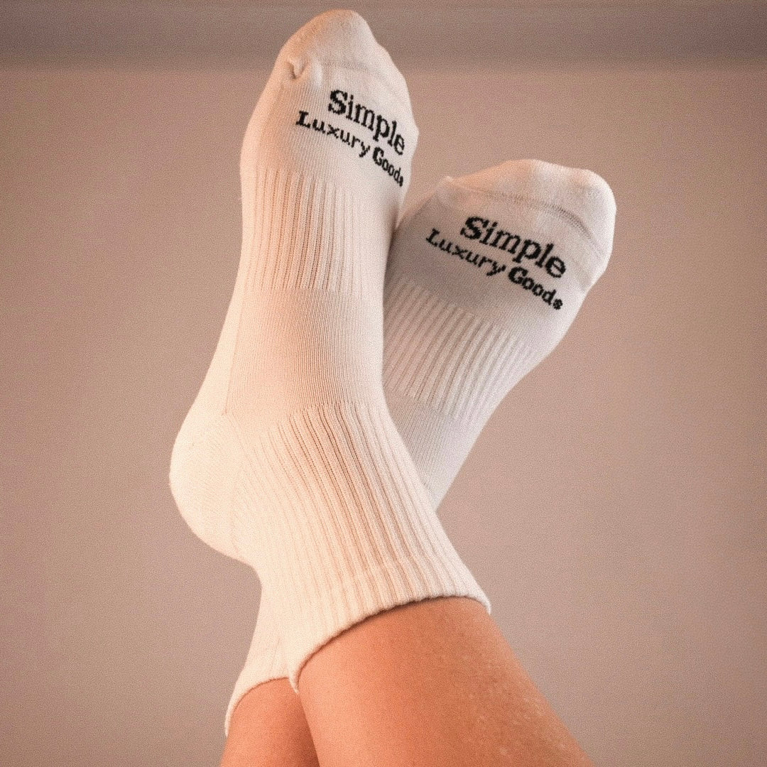A pair of white ribbed quarter socks worn on feet, with the words 'Simple Luxury Goods' jacquard knitted in black on the toes, shown against a plain beige background.