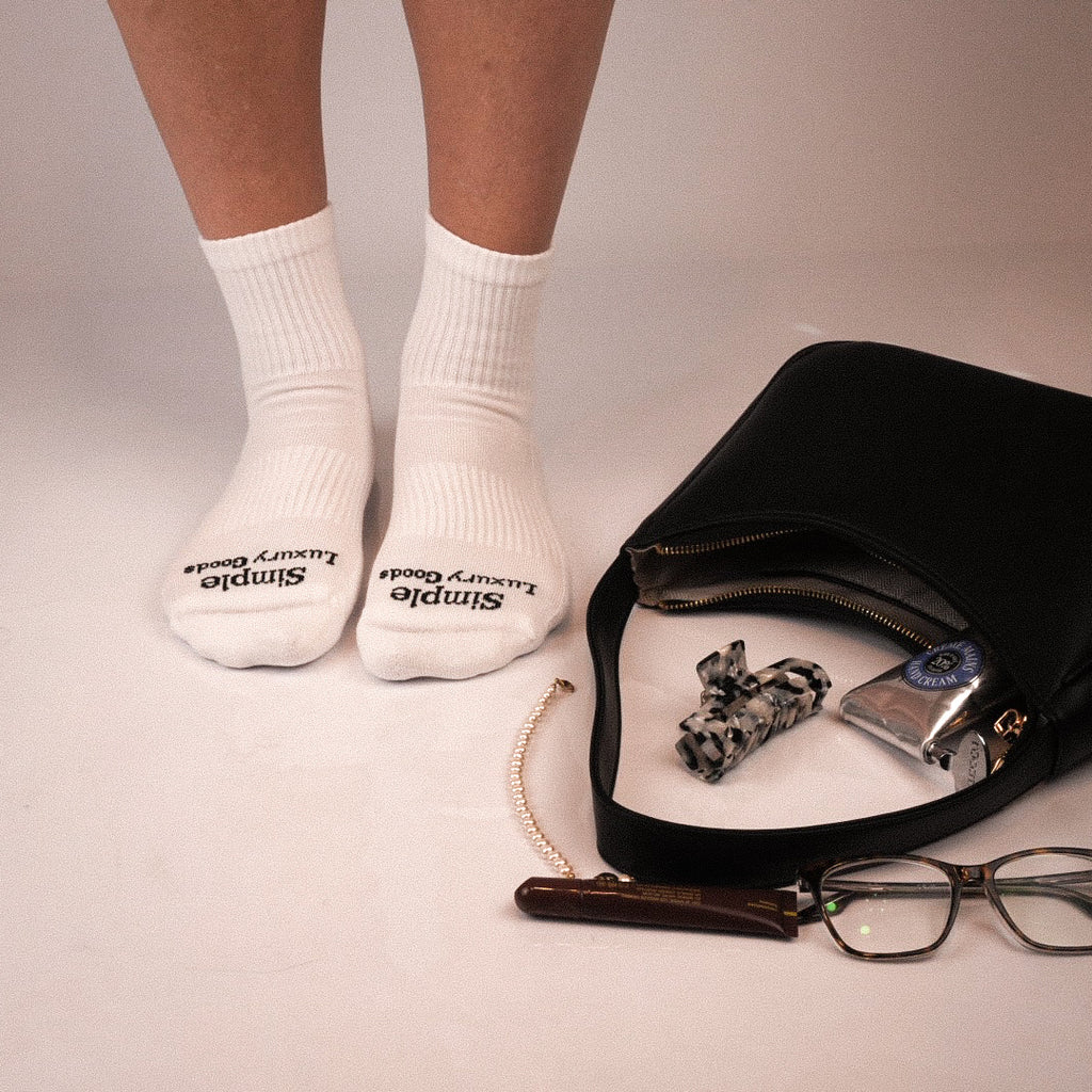 White quarter socks with black 'Simple Luxury Goods' branding on the toes, worn on a person's feet with a black bag, eyeglasses, and accessories nearby on a light beige studio background.