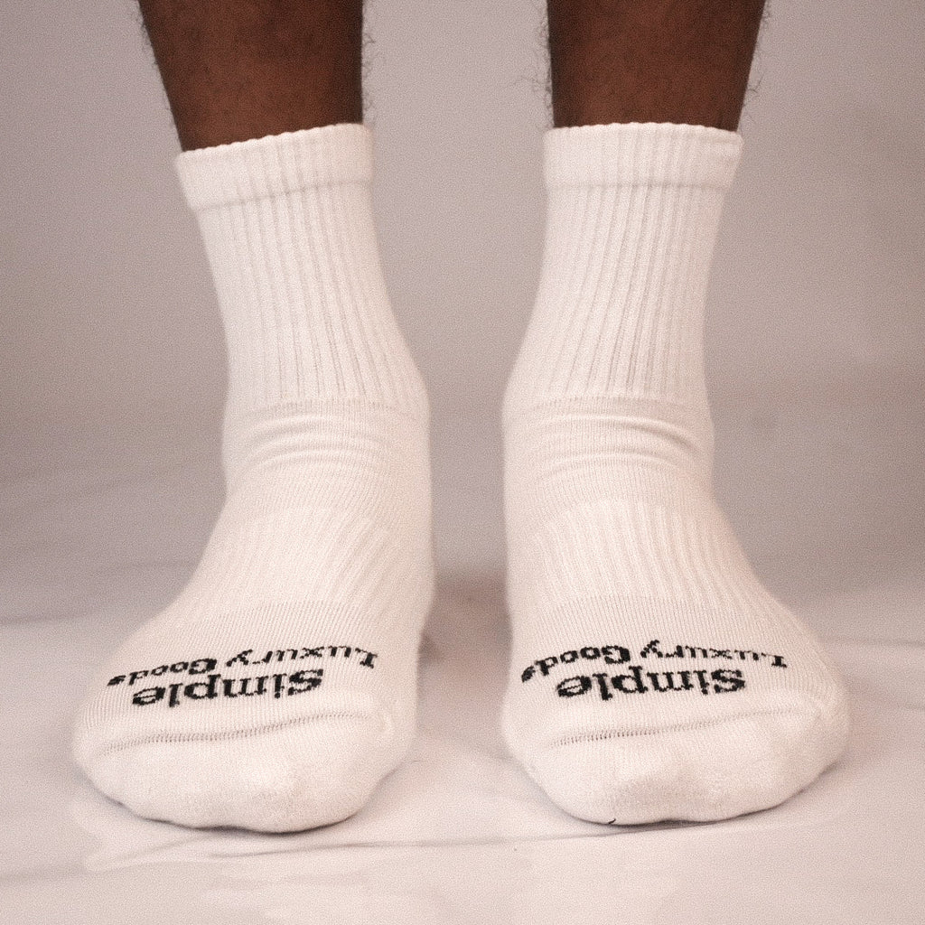 A pair of white ribbed quarter socks worn on feet, with black text reading 'Simple Luxury Goods' jacquard knitted on the toes, shown against a light gray background.