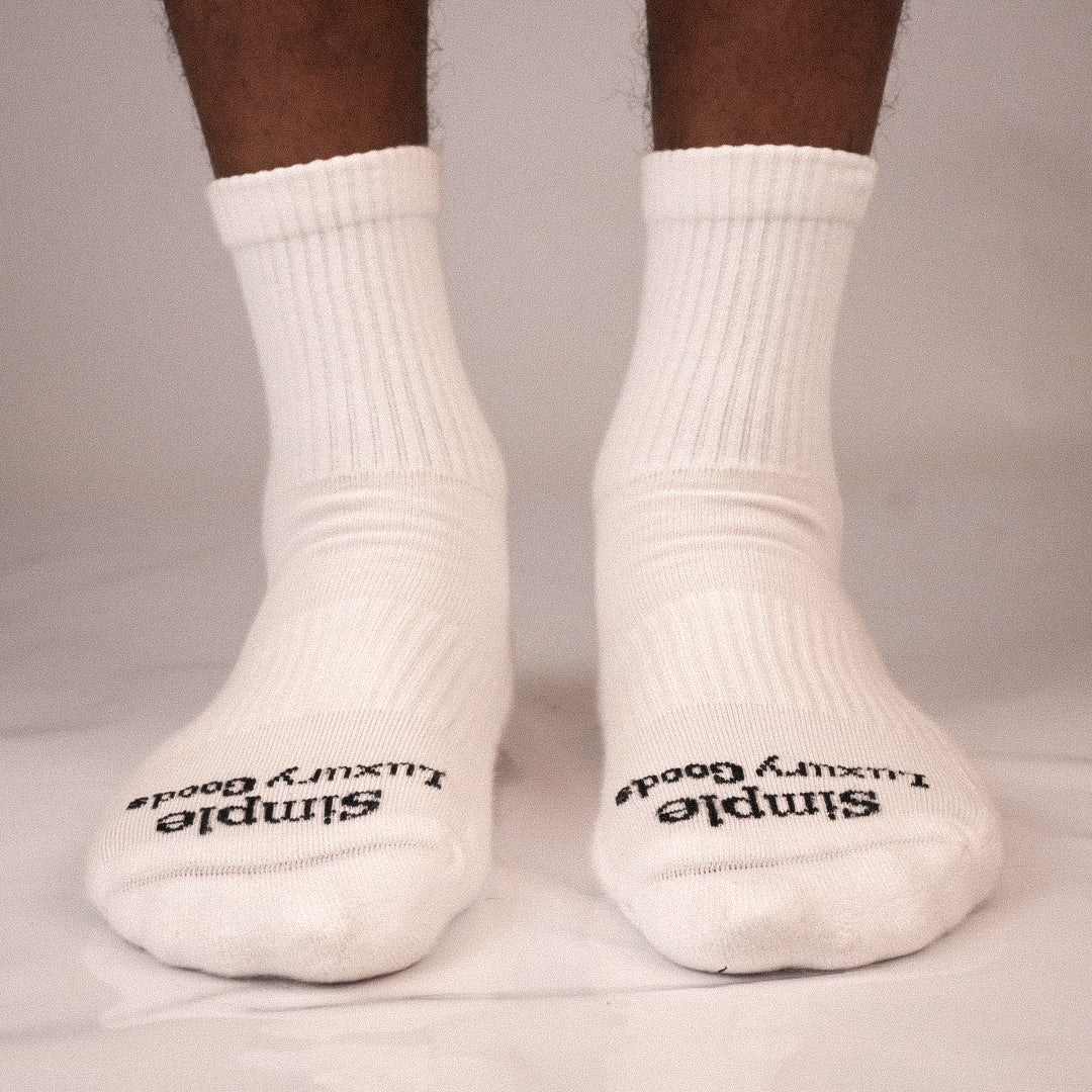 A pair of white ribbed quarter socks worn on feet, with black text reading 'Simple Luxury Goods' jacquard knitted on the toes, shown against a light gray background.