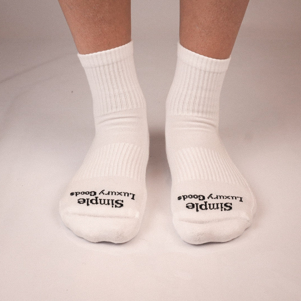 A pair of white quarter socks worn on feet, with black jacquard knitted text reading 'Simple Luxury Goods' on the toes, shown against a plain light background.
