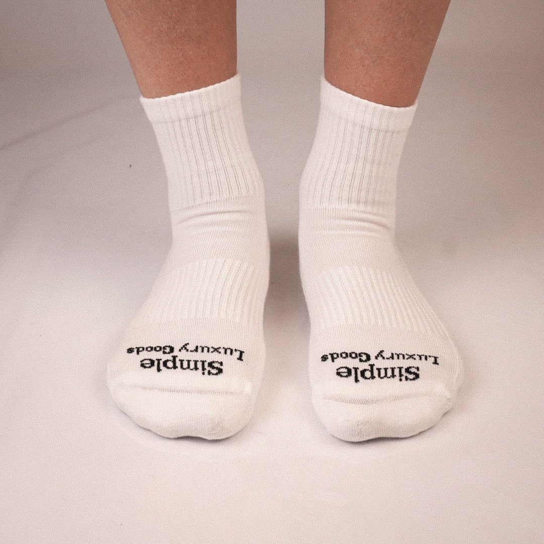 A pair of white quarter socks worn on feet, with black jacquard knitted text reading 'Simple Luxury Goods' on the toes, shown against a plain light background.