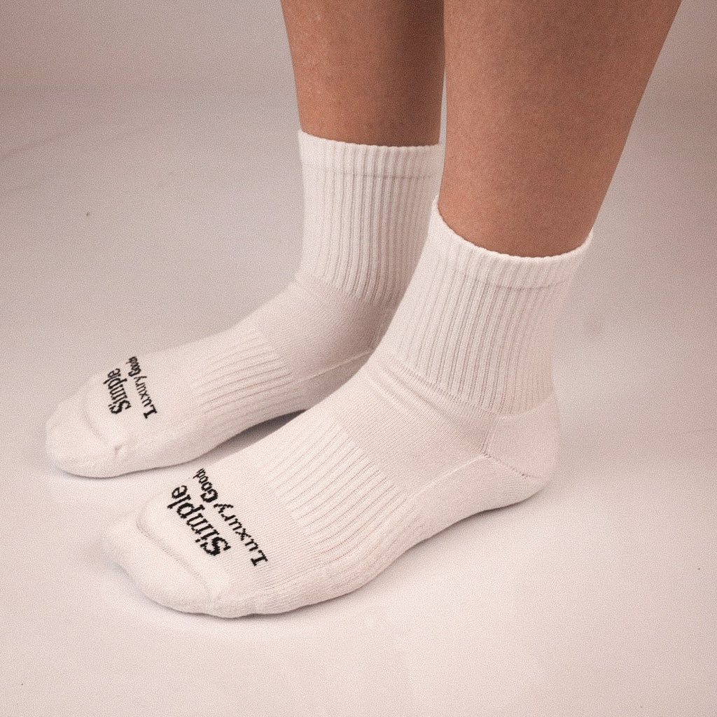 A pair of white ribbed quarter socks worn on a person's feet, with black text reading 'Simple Luxury Goods' jacquard knitted on the toes.